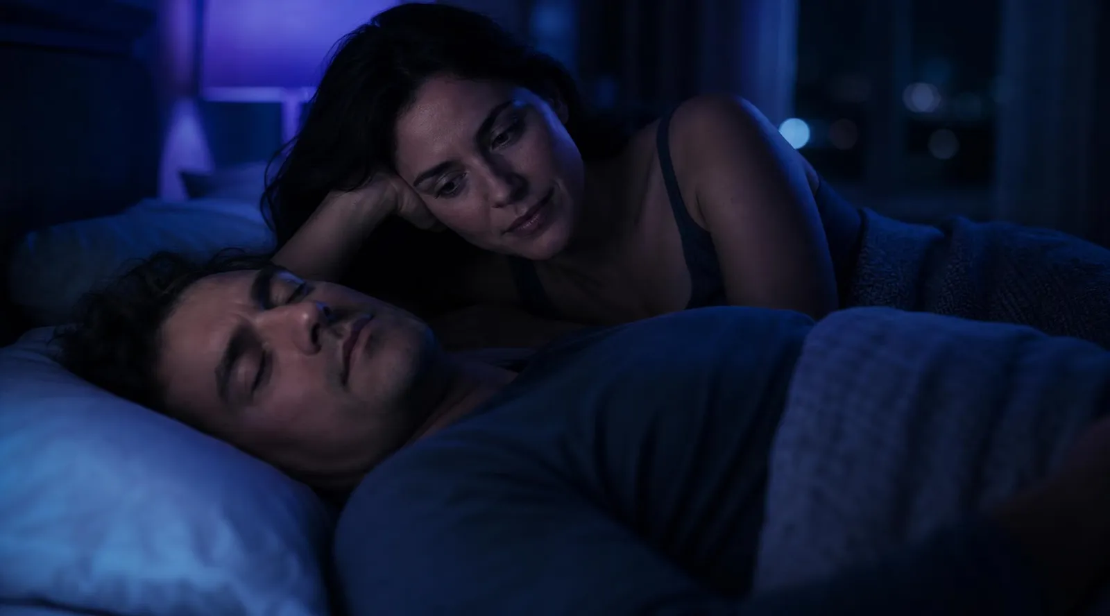 A couple in bed at night in a blue-lit bedroom, one partner asleep while the other lies awake beside them, illustrating how snoring keeps couples from resting — and why a snoring app built for couples measures both people.