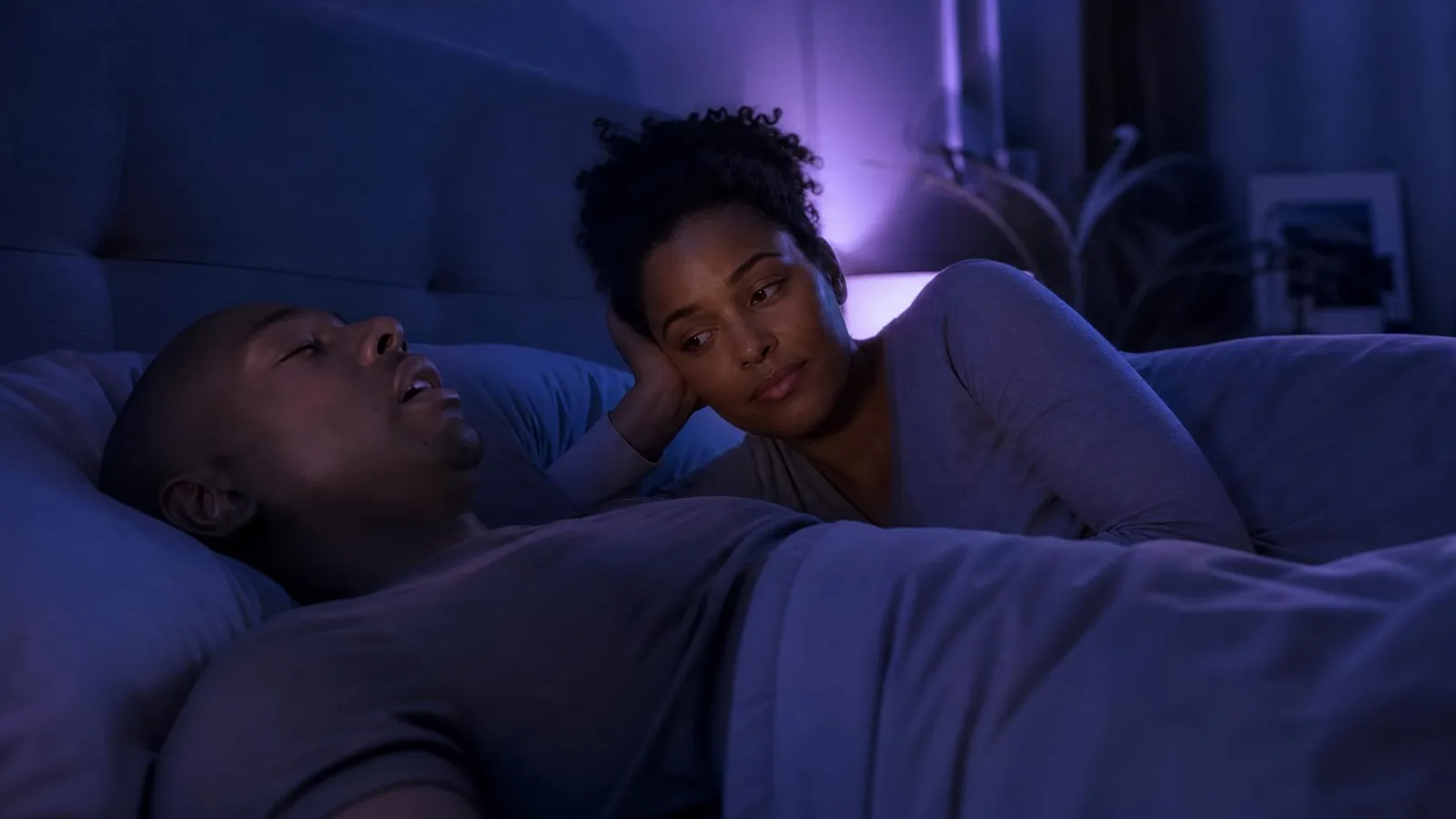 A man sleeping on his back with his mouth open while his partner lies awake beside him, watching with concern — the bed-partner who notices breathing pauses, central to STOP-BANG sleep apnea screening.